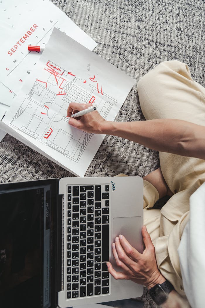 Overhead view of a person sketching floor plans next to a laptop on a carpet.
